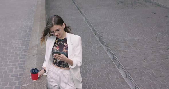 Happy Girl Holds a Cup of Coffee and Uses Smartphone with Smile in Windy Day