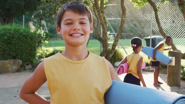 Portrait of happy caucasian schoolboy holding yoga mat after yoga lesson outdoors