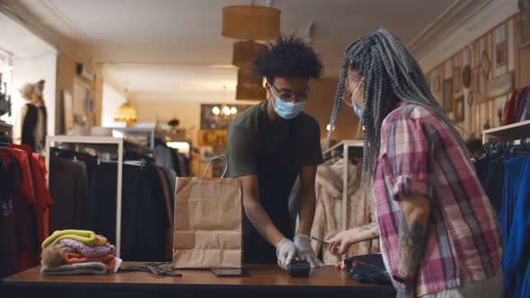 Young Woman in Safety Mask Paying with Credit Card for Purchase in Clothes Store