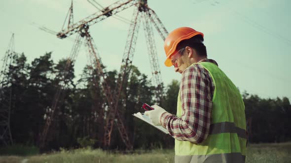 Caucasian Engineer in Protective Uniform and Orange Hard Hat Holds Walkietalkie and Clipboard on