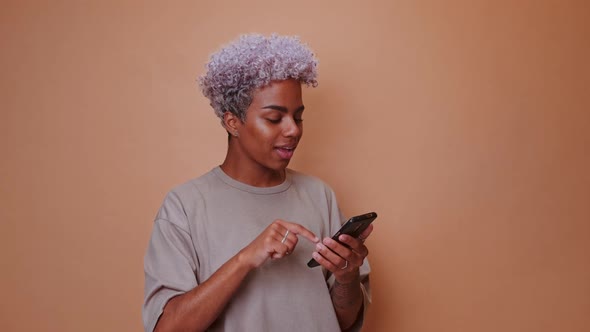 Young African American Woman with Mobile Phone in Hand Stands in Brown Studio