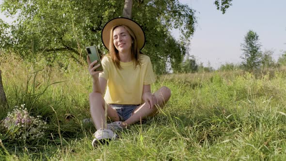 Smiling Talking Woman Sitting on Meadow Looking at Smartphone Screen Using Video Call
