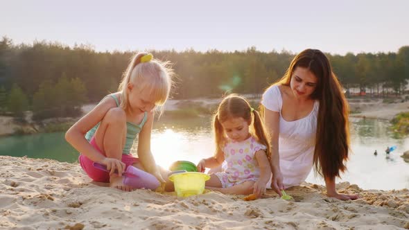 Young Mother Plays in the Sand with Two Daughters. At Sunset on the Background of the Beautiful Lake