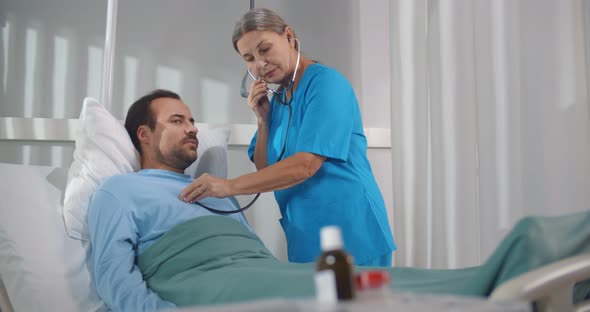 Senior Female Doctor Using Stethoscope To Check Heart Rhythm of Male Patient Lying in Hospital Bed