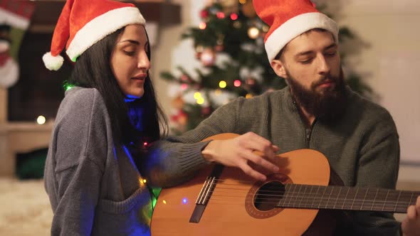 Portrait of Happy Couple in Santa's Hats Singing Songs at Home. Man Playing Guitar. Woman with