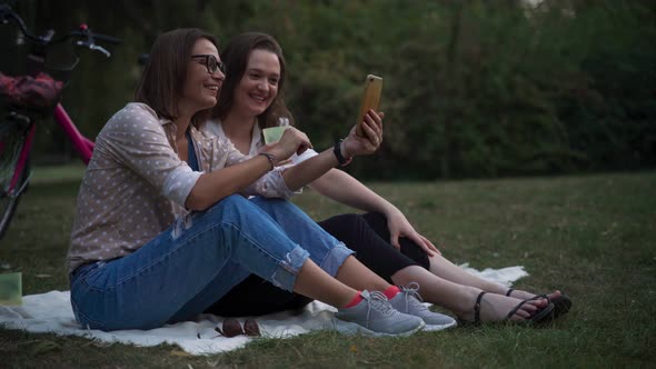 Two Young Cheerful Women Sitting at the Park and Taking a Video Call