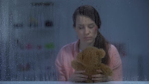 Hopeless Woman With Wounded Face Hugging Teddy Bear Behind Rainy Window, Support