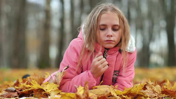 A Little Girl Indifferently Lies on the Autumn Foliage in a City Park