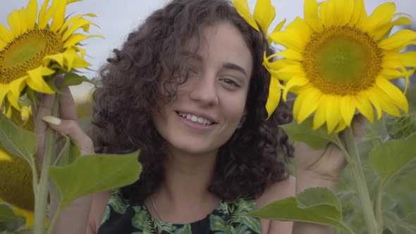 Portrait of Cute Curly Girl Looking at the Camera Smiling Covering Her Face with Two Sunflowers