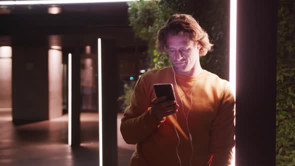 Caucasian male wearing earphones, smiling, lying on a wall, using his phone in the evening