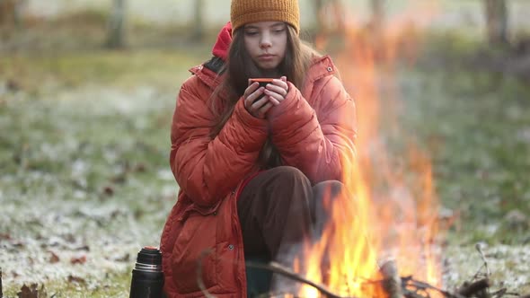 Teen girl with mug sitting near bonfire in camp