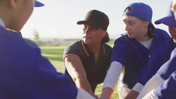 Diverse group of female baseball players and coach on pitch, in a huddle, stacking hands