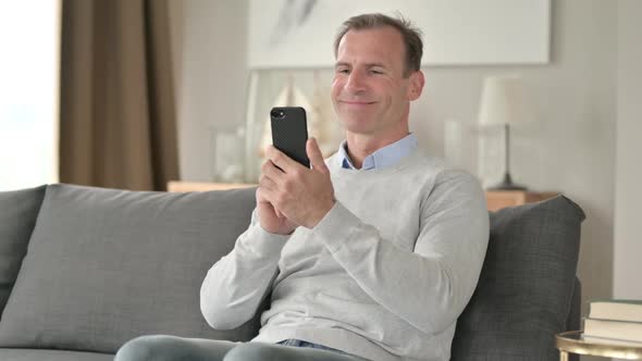 Middle Aged Businessman Celebrating Success on Smartphone on Sofa