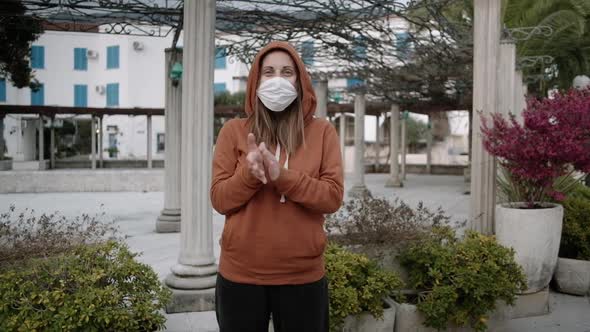 Woman in orange sweater and medical mask stay  and claps on empty street.