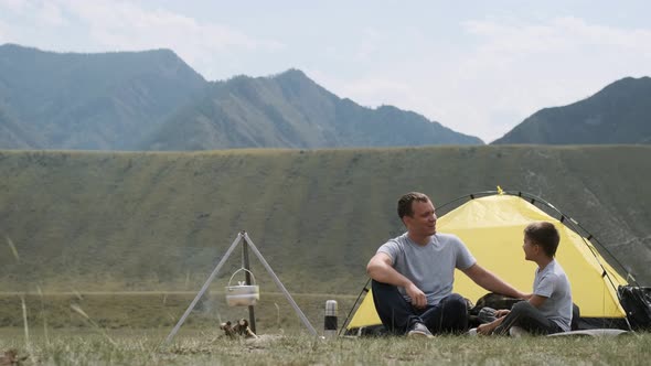 Happy Father and Son Are Sitting Near the Tent and Bonfire and Laughing