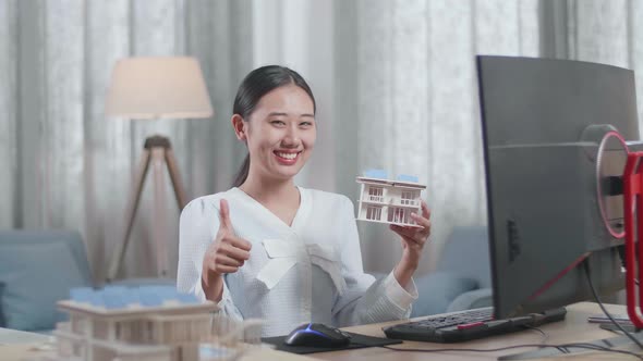 Smiling Asian Woman Engineer Holding House Model Showing Thumbs Up Gesture To The Camera