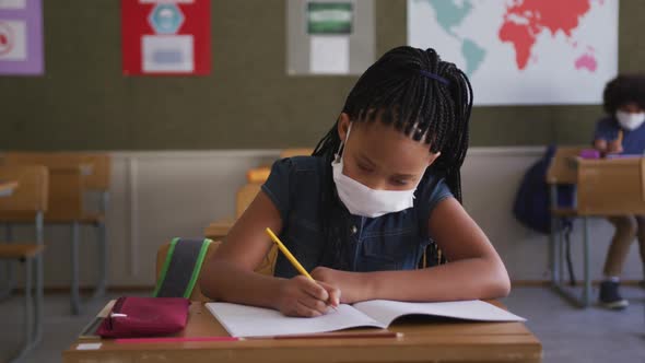 Girl wearing face mask writing while sitting on her desk at school 