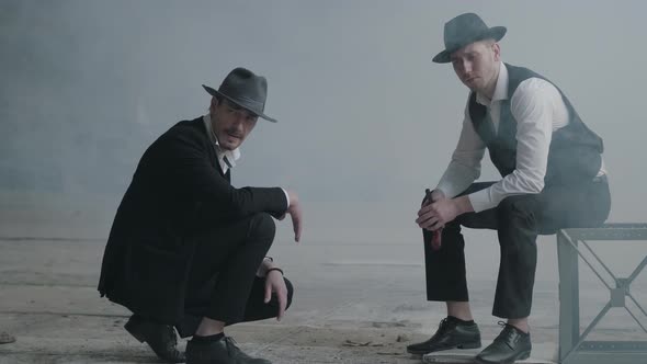 Two Stylish Men in Hats, One Sitting on the Chair, Another Squatting in an Abandoned Building