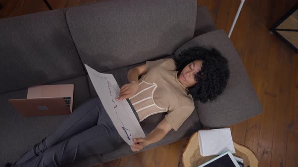 Top View Wide Shot of Serious Professional Business Analyst Examining Graph Lying on Couch in Home