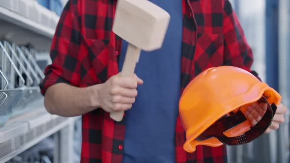 Unrecognizable Young Man Hitting Hard Hat with Hammer Standing at Shelves in Hardware Store