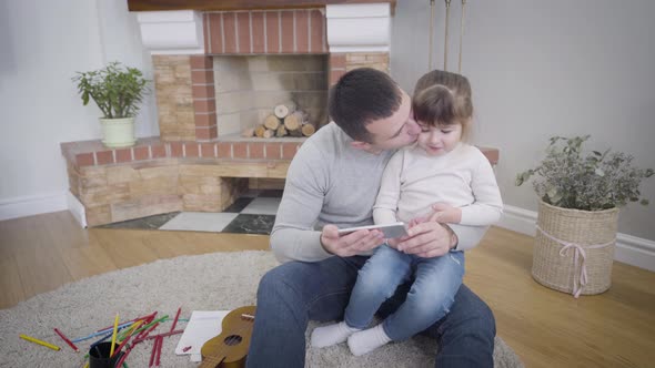 Portrait of Cute Caucasian Little Girl Sitting with Father and Watching Cartoons on Smartphone