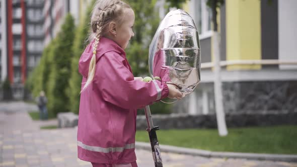 Side View of Cheerful Girl with Pigtails Riding Scooter. Happy Pretty Caucasian Child with Balloon