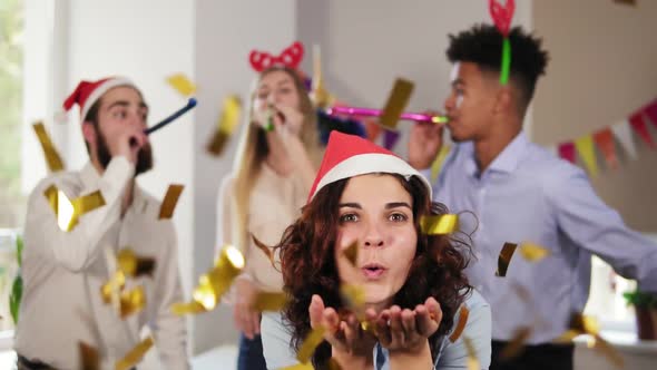 Young Attractive Woman Blowing Golden Confetti From Hands Wearing Christmas Hat While Her Coworkers