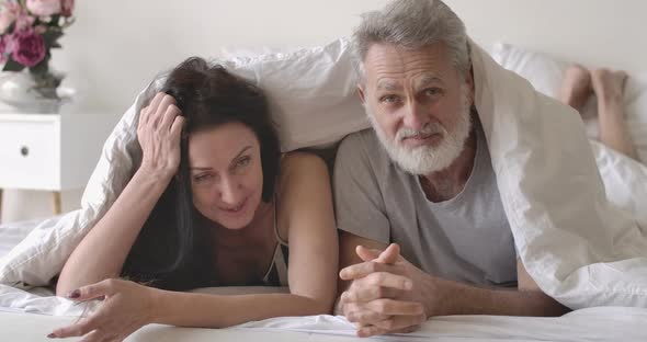 Positive Mature Caucasian Couple Lying in Bed Under Blanket, Talking and Laughing. Happy Senior Man