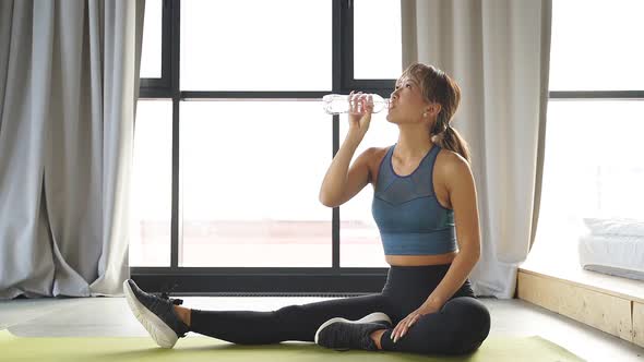 Young Japanese Woman After Fitness Classes Sitting on a Fitness Mat Drinks Water From a Bottle