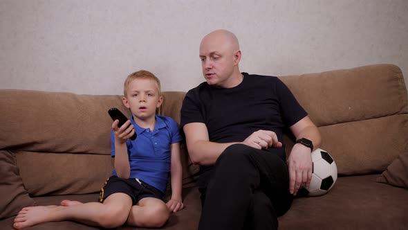 Father and Son on the Couch with a Remote Control and a Ball in Front of the TV