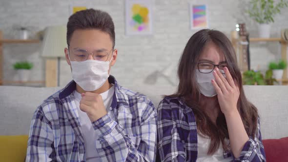 Sick of a Cold Young Sian Couple Sitting in Protective Medical Masks on the Couch