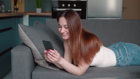 Young Woman Relaxing on Sofa Texting Using Smartphone Browsing Online Playing Mobile Games Enjoying