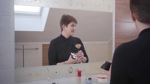 Portrait of Senior Beautiful Caucasian Woman Looking at Big Mirror and Applying Face Powder