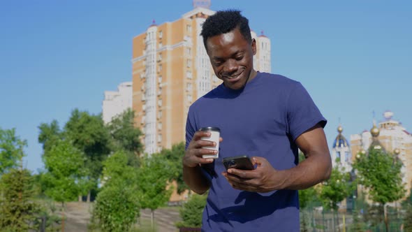 Smiling Afroamerican Man Walks Street Uses Mobile Phone Holding Cup with Coffee