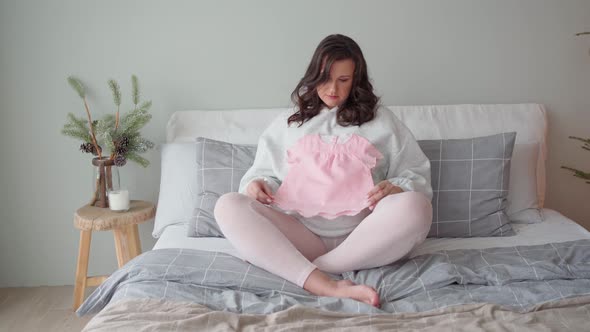 a Young Beautiful Pregnant Woman Sits on the Bed and Examines a Pink Pink Little Dress for Her