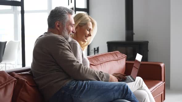 Cheerful Middleaged Couple with a Laptop Sitting on the Comfortable Leather Couch
