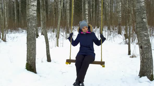 A young caucasian girl swinging on winter forest in snow covered winter birch forest at sunny day, a