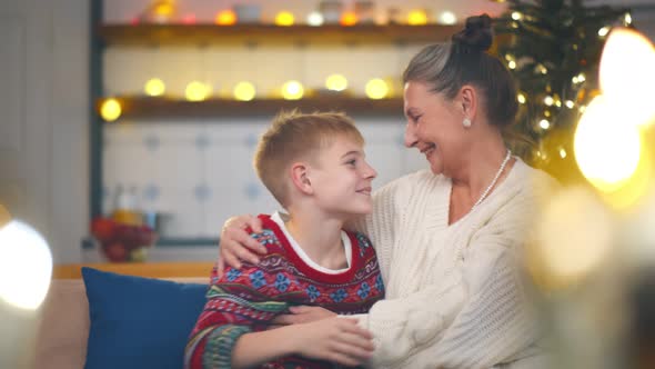 Happy Grandson Sitting with Grandma on Couch and Embracing on Christmas Eve