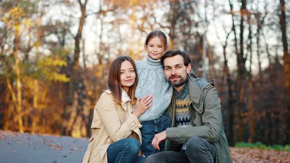 Young Smiling Mother and Father Sitting Squatting in Autumn Park and Hugging Their Little Daughter