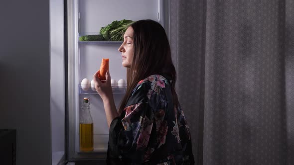 Lady Chooses Cake Over Carrot Near Fridge in Dark Kitchen