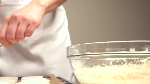 Pastry maker passing cupcake mix to molds from bowl with scoop and spoon