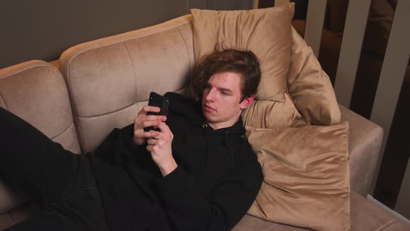 Smiling Happy Young Man Use Phone Read Good News Lying on Sofa at Home