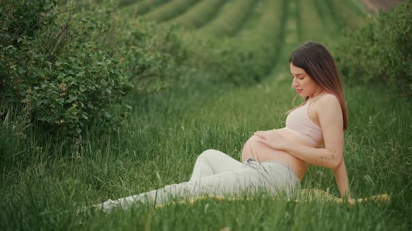 Pregnant young woman with dark hair in pink top and light pants sitting on green grass in field