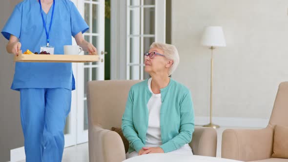 Caretaker carries tray with breakfast and hugs positive single mature lady