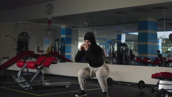 Wide Shot Portrait of Confident Slim Sportive Middle Eastern Woman in Hijab Doing Squats in Gym