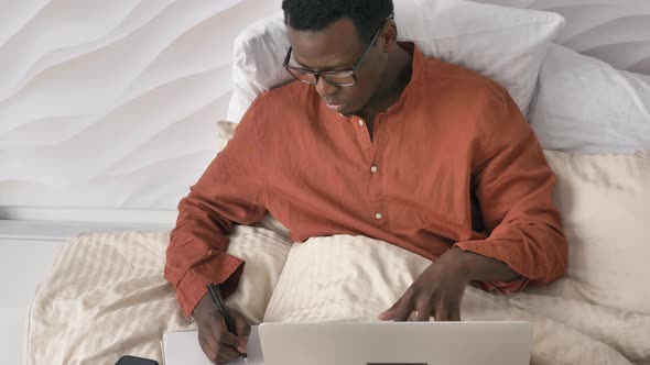 AfroAmerican Man with Glasses Makes Notes Working on Laptop