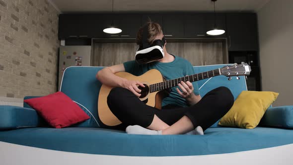 Young Man Sitting on Sofa with the Virtual Reality Glasses Is Playing at the Guitar