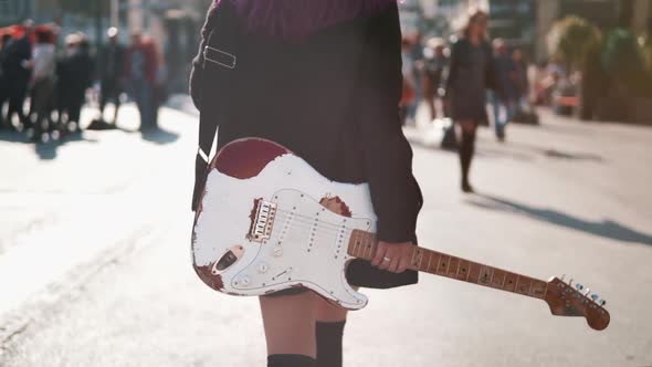 Daring Rebel Woman with Purple Hair Walking with Electric Guitar at City Center During Bright Sunset