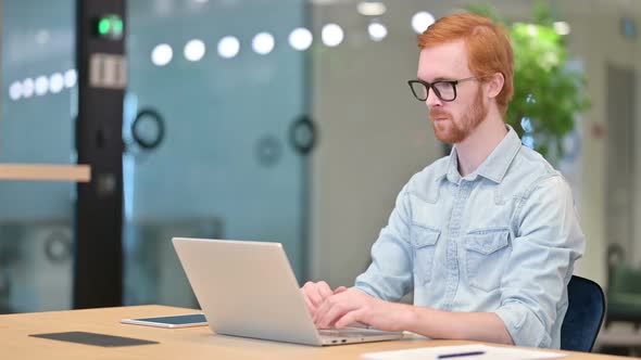 Focused Young Redhead Man Working on Laptop in Office