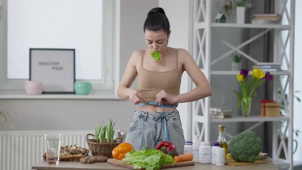 Slim Fit Caucasian Woman Measuring Waist Chewing Salad Leaf Standing at Home Indoors